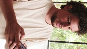 Vertical video, Smiling young man with curly hair and beard using mobile phone while standing in cozy living room