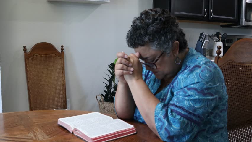 Middle-age Hispanic Woman praying at kitchen table with open bible