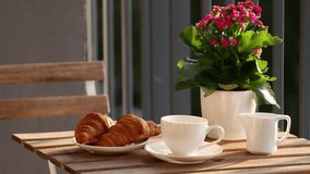 Female hands in a white sweater pour tea from a white teapot into a white ceramic cup outside, a table served with croissants and flowers.