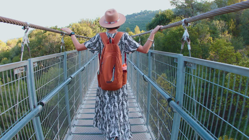 happy laughing woman in a hat walks along a pedestrian suspension bridge over the river on a sunny autumn day. back view.