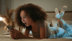 African American woman happy shocked young girl mobile phone gadget user lying on bed in home bedroom amazed astonished female winning smartphone victory wonder achievement night scrolling good news - Powered by Shutterstock - Get 15% off with code: PIKWIZARD15