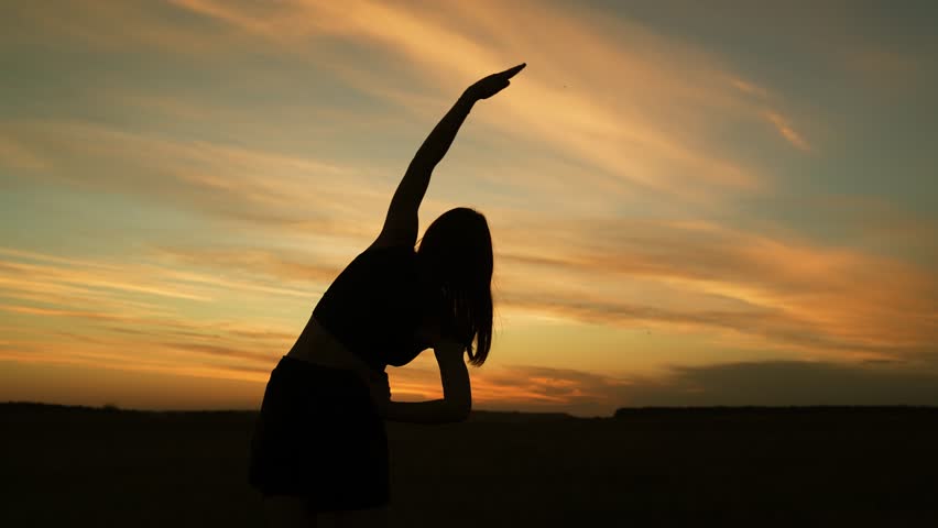 Silhouette of flexible woman doing side bends in evening park. Woman athlete exercises alone in empty sunset field. Active sportswoman trains standing in meadow at back cloudy orange sunset light