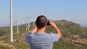 A young man shooting landscape video, photos for social media on a smartphone. A man takes horizontal panorama of wind turbines on a mountainous landscape using his mobile phone.