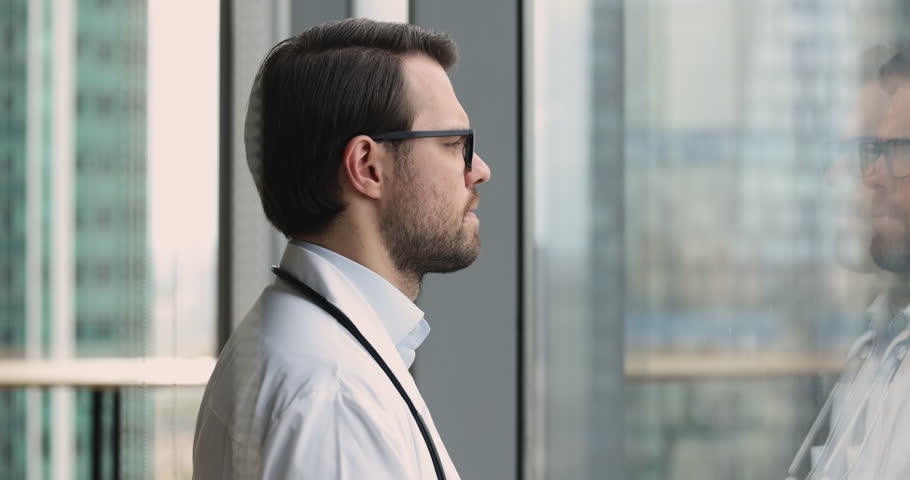 Close up young 35s desperate or fatigued male therapist in white uniform stands near window with bowed head feels concerned, looks terribly upset due to medical error, overloaded. Professional failure Royalty-Free Stock Footage #1109929433