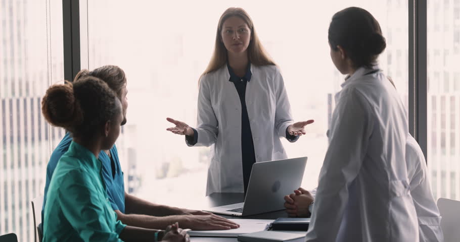 Multinational team of therapists in diverse uniforms listen speech of female colleague, head physician gathered together in modern clinic conference room. Medical concilium event, meeting of GP staff Royalty-Free Stock Footage #1109929447