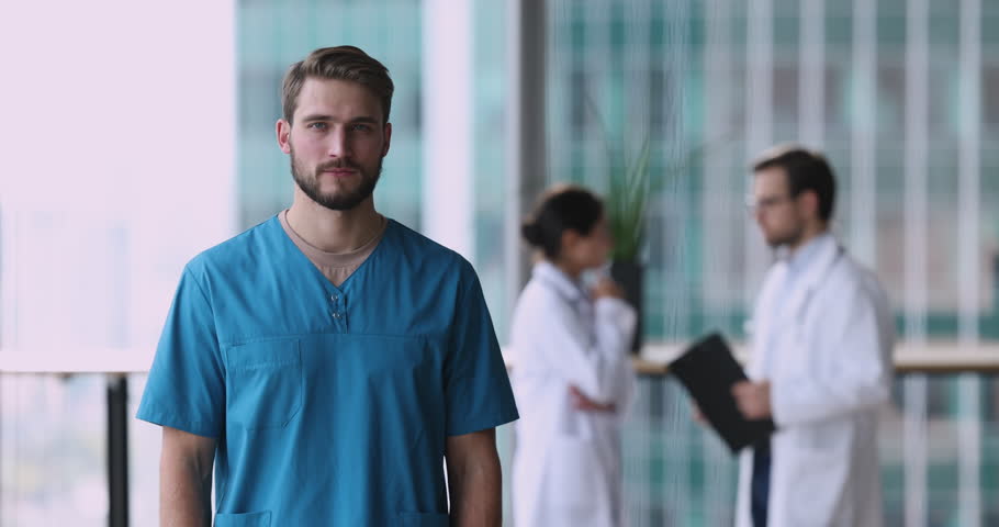 Handsome positive male physician GP wear blue uniform pose in modern clinic office smile feels satisfied with career. Professional medical services, vocation, profession, reliable physician portrait Royalty-Free Stock Footage #1109930297