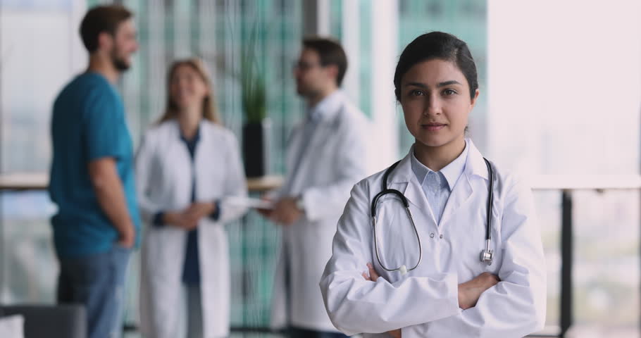 Indian female general practitioner in white medical coat smile look at camera pose in clinic office with doctors on background. Professional physician portrait, medicine, hospital services, medicare Royalty-Free Stock Footage #1109930323
