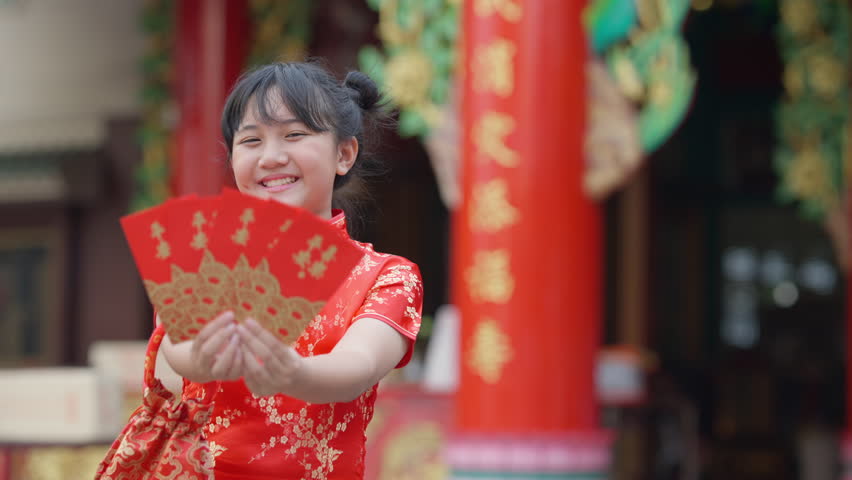 Portrait of Happy Little Asian girl in Chinese red dress holding red envelopes in front of in Chinese temple shrine. Chinese lunar new year festival, holiday celebration, religion meditation concept.