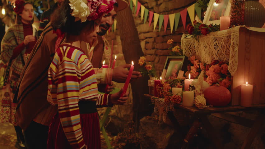 Medium shot of little Mexican girl with flower wreath in her hair lighting candle while standing by altar of her grandmother with other family members, celebrating Dia de Muertos together in backyard Royalty-Free Stock Footage #1109943783