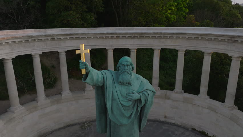 Pull back footage of historic tourist monument with St. Gellert statue in park. Revealing trees and paths. Cars driving on road around. Budapest, Hungary