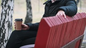 A young woman drinks coffee from a paper cup on a bench in an autumn park. Female hands are holding a cup of aromatic coffee. A woman enjoying life in slow motion. Autumn season.