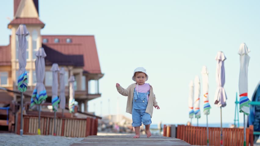 Small child joyfully runs along wooden path on sandy beach past bright beach umbrellas. Toddler girl passes large house imitating fortress