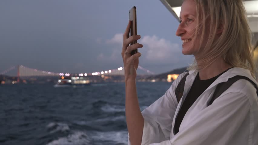 Young woman taking pictures of sea and coastal city while traveling on water by boat or ferry in evening or at night. Lights of city are burning. Using camera on your smartphone during boat cruise