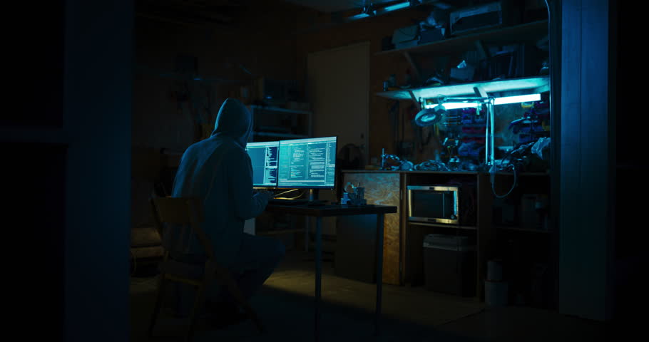 Home Garage Late At Night: Anonymous Male Hacker Wearing Hoodie Breaks into Data Servers, DDOS Attack, Phishing Attack, Malware. Man Writing Code For Trojan Horse Virus. Darknet Cyber Crime Concept.