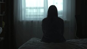 Back view Silhouette of a millennial woman sitting on a bed in the dark bedroom, depression, pain. Portrait of Unhappy sad woman sitting against window and thinking about her life - Powered by Shutterstock - Get 15% off with code: PIKWIZARD15