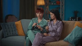 Medium-full video capturing two young women wearing pajamas sitting on the couch in a room, gossiping, talking about something as they paint each other's nails.