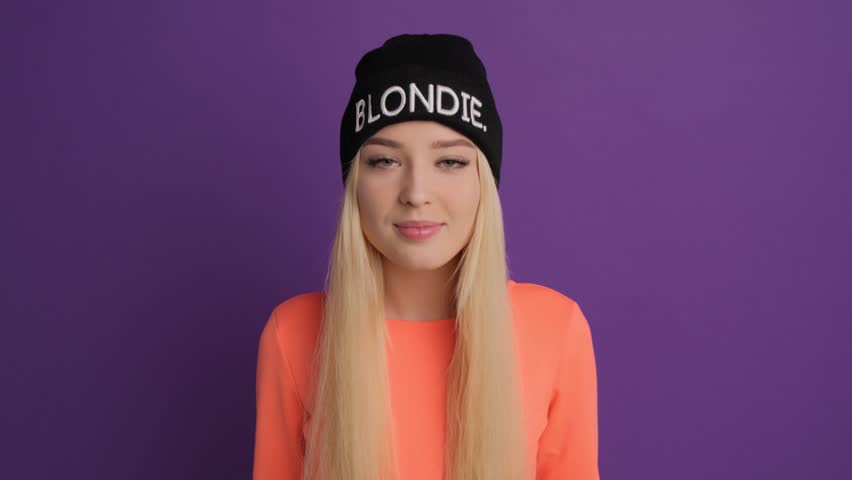A blonde woman in a black cap adjusts her hat and smiles.