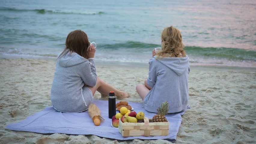 Bundled up women in autumn attire, clutching warm cups in their hands, sipping hot drink on sea beach during evening. Having picnic on summer evening