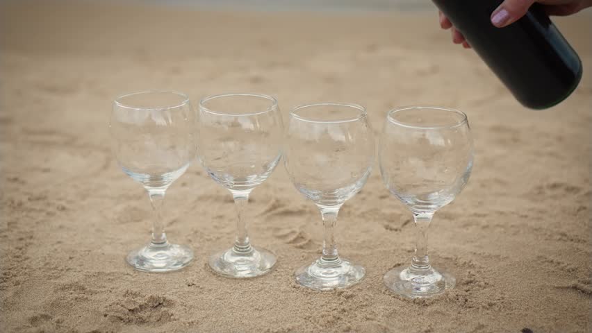 Close up woman hand Pouring red wine in four glasses standing in row on sand of sea beach with waves in background. Alcohol drinking, enjoying life, active rest at seashore