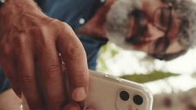 Vertical video, Close-up of hands of an elderly man using phone while sitting in park, background blurred