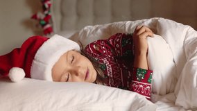 Little child girl wearing red Christmas pajamas and white fluffy Santa hat lie pillow under cozy warm blanket. Sleep at night and smile sweet dreams. Merry Christmas Happy New Year Holiday 2024 - Powered by Shutterstock - Get 15% off with code: PIKWIZARD15