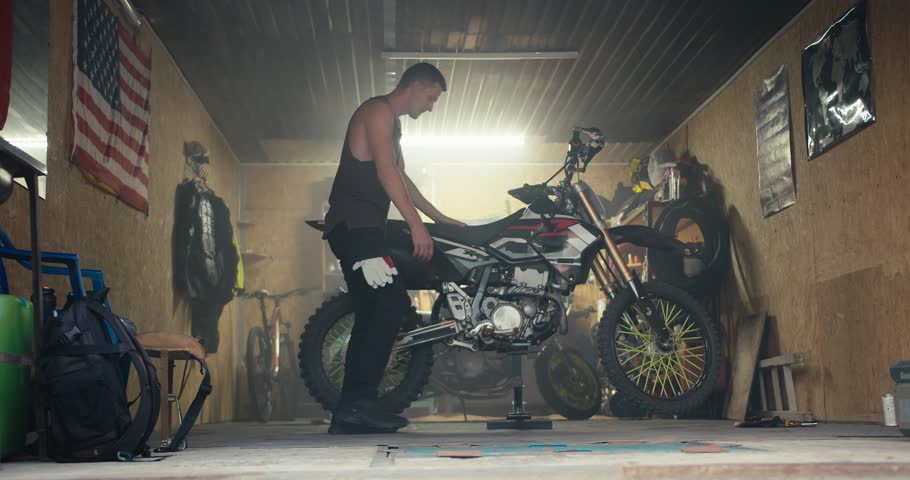 A male mechanic with a short haircut in a gray T-shirt removes his bike from the jack and takes it out of his garage workshop. Male mechanic completing motorcycle repair Royalty-Free Stock Footage #1110000291