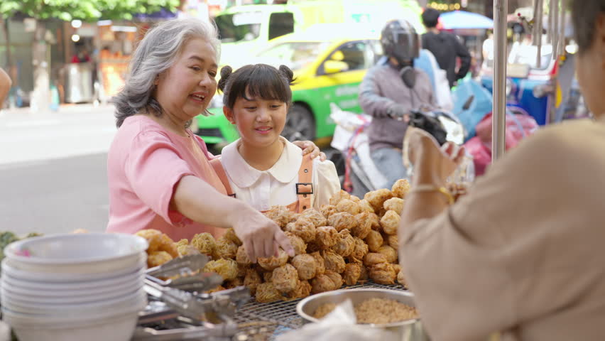 Asian family grandmother and grandchild girl buying and eating street food together at street market. Senior woman and little girl enjoy outdoor lifestyle travel in the city on summer holiday vacation