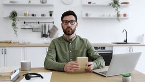Caucasian bearded man in eyeglasses touching chin and looking at small cardboard parcel with focused facial expression. Social media influencer streaming unpacking process of online order from home.