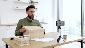 Happy male blogger in casual wear unpacking box with new wireless laptop and recording video using modern smartphone with tripod. Concept of online shopping, social media and technology.