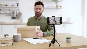 Caucasian bearded man making surprised facial expression while unpacking cardboard box during live streaming. Male influencer using modern smartphone and tripod for creating new content.