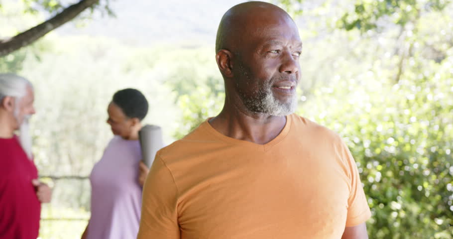 Happy african merican senior man holding yoga mat, with freinds in sunny nature, slow motion. Summer, retirement, yoga, wellbeing and healthy senior lifestyle, unaltered.