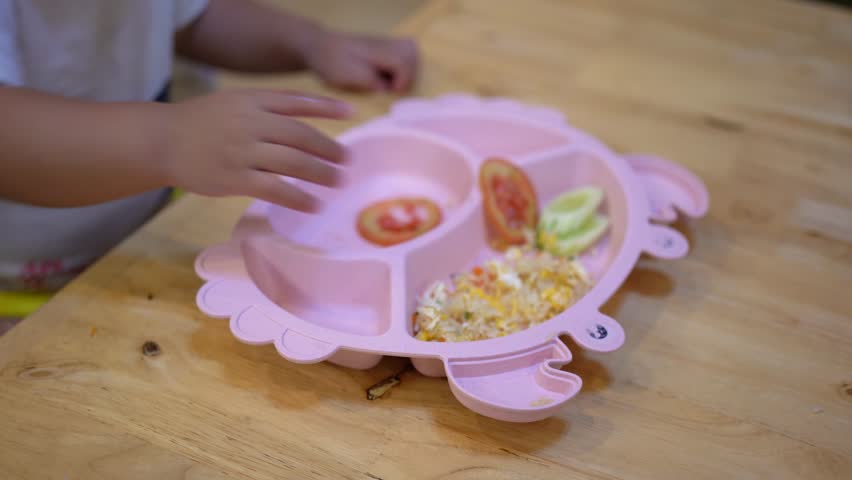 Acion of a baby is using hand for eating meal in the pink plastic dish which is plated on wooden table. Baby lifetyle and learning practice, 4k footage.