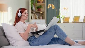 modern technology, a young woman using a headset talking on a video call on a laptop while lying on sofa in room, looking at camera