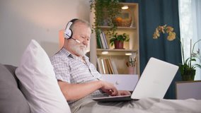 modern old man using microphone and headphones talking on video call on laptop while relaxing at home