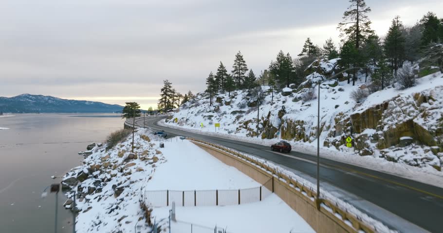 An aerial of a red car driving on the road alongside the snowy hill and frozen lake