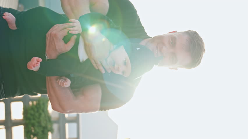 Strong Caucasian man holding a baby wearing black suit. Infant boy starts to cry and dad waves calming him. Sun at backdrop. Vertical screen.