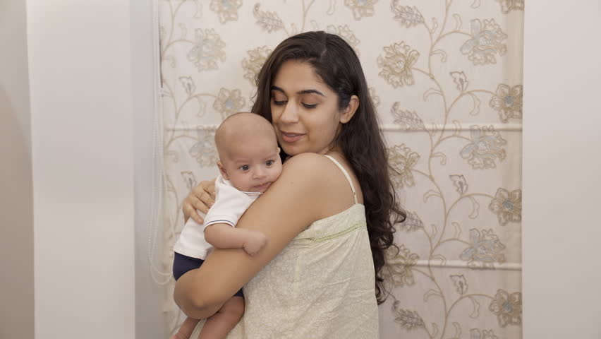An attractive female petting a baby to make him sleep - caring mother, new mother, mother-baby bonding, happy parenting. Pretty Indian lady holding a newborn child in her arms while cuddling him - ...