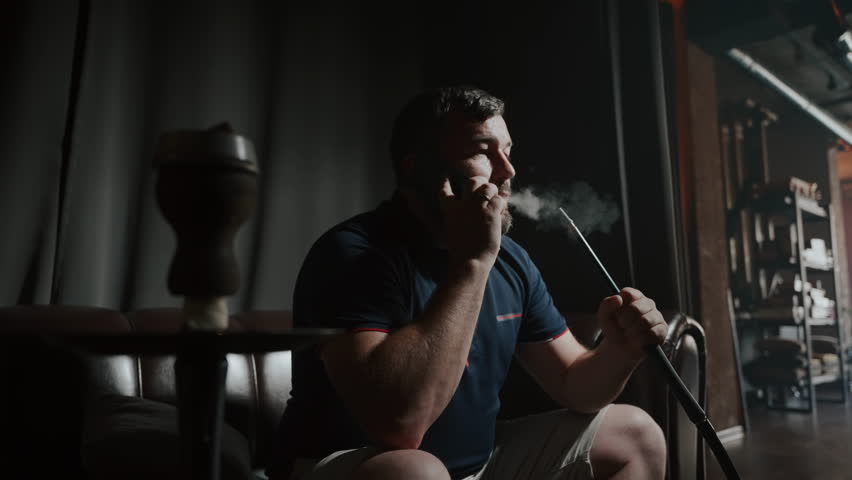 Business businessman exhaling smoke while listening to conversation on phone. Slow motion, low key, relaxing in a hookah lounge.