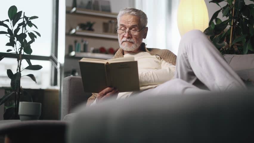Portrait of gray haired senior man under the blanket reading book literature at cozy home Calm relaxed mature male with glasses enjoying weekend leisure time alone indoors Royalty-Free Stock Footage #1110043709