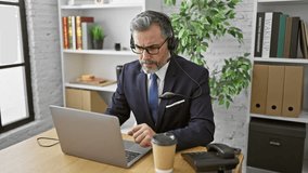 Attractive grey-haired young hispanic man mastering the office call center game, video chatting with client on laptop, adorned in suit and tie, listening to cool tunes.