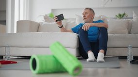 Senior man finds video training in smartphone for doing sport exercises at home sits on yoga mat in living room. Elderly male in sportswear prepares to workout. Sport active healthy lifestyle concept.