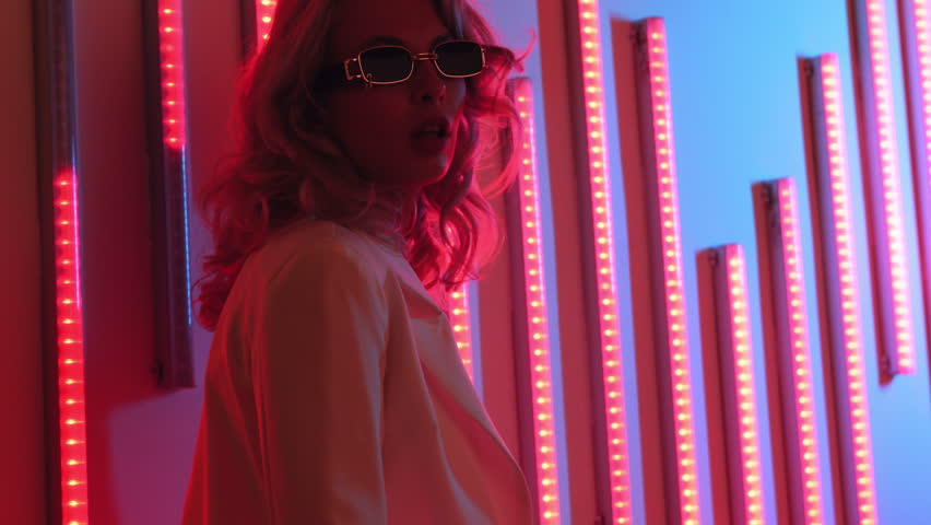 Young beautiful hipster woman in street style fashion concept. Hot model wearing white suit and sunglasses. Female posing in neon light in studio interior
