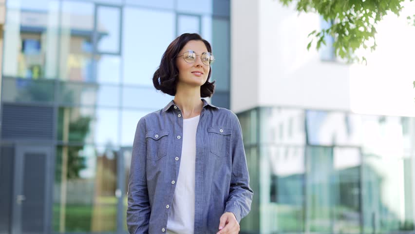 Portrait of smiling female student looking at camera while standing in campus space near university building. Head shot of a positive brunette teacher in glasses and a shirt posing with crossed arms Royalty-Free Stock Footage #1110050621