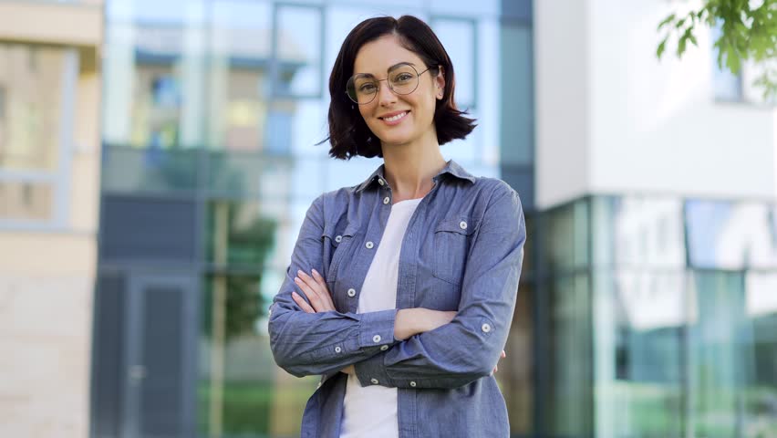 Portrait of smiling female student looking at camera while standing in campus space near university building. Head shot of a positive brunette teacher in glasses and a shirt posing with crossed arms