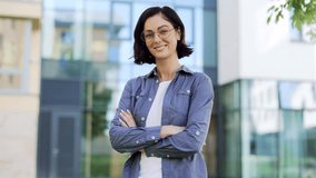 Portrait of smiling female student looking at camera while standing in campus space near university building. Head shot of a positive brunette teacher in glasses and a shirt posing with crossed arms - Powered by Shutterstock - Get 15% off with code: PIKWIZARD15