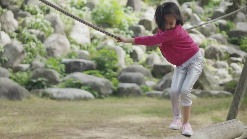Asian kid girl, alone in her summer activity with playful exploration and determination by walking onto a wooden balance beam with holding rope in beautiful forest park in summer sunlight. Royalty-Free Stock Footage #1110065837