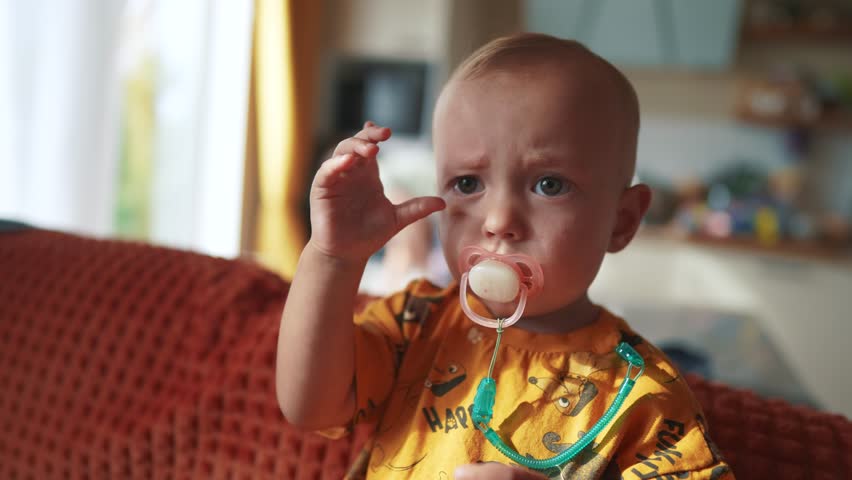 a small newborn sits on the couch with a pacifier in his mouth. happy family childhood dream concept. a little child with a pink pacifier sits and looks lifestyle at the camera baby face close-up Royalty-Free Stock Footage #1110066221