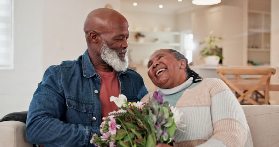 Sofa, flowers and love with a senior black couple in their home living room for anniversary celebration. Birthday, valentines day and an elderly husband hugging his wife while laughing for romance Royalty-Free Stock Footage #1110072735