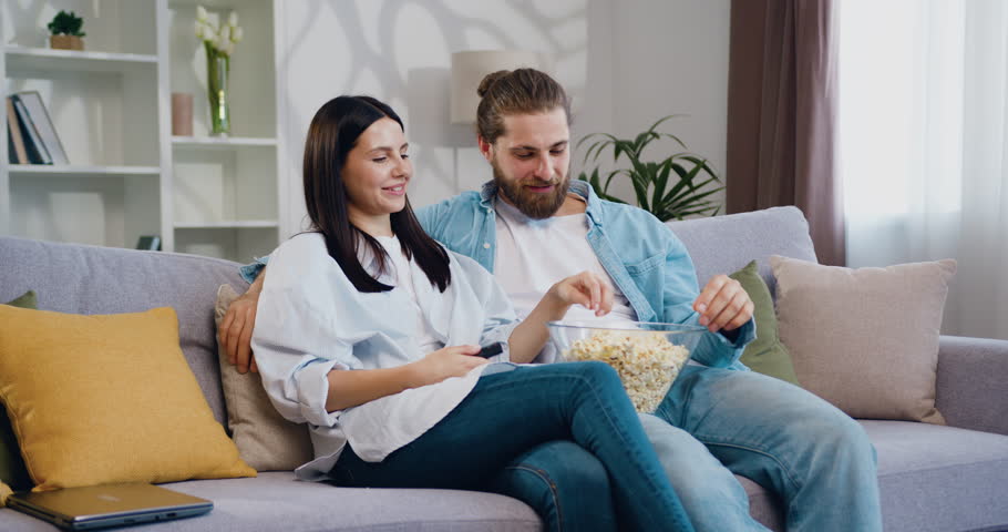 Attractive carefree couple man and woman sitting on sofa in front of TV ,eating popcorn, talking and bouncing watching their favorite series on couch after hard day. Young family eating food Royalty-Free Stock Footage #1110075253