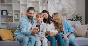 Happy family adult parents with cute school kids children using smartphone apps laughing having video call with technology together siting on sofa at home. Mother Father preschool kids hold look at